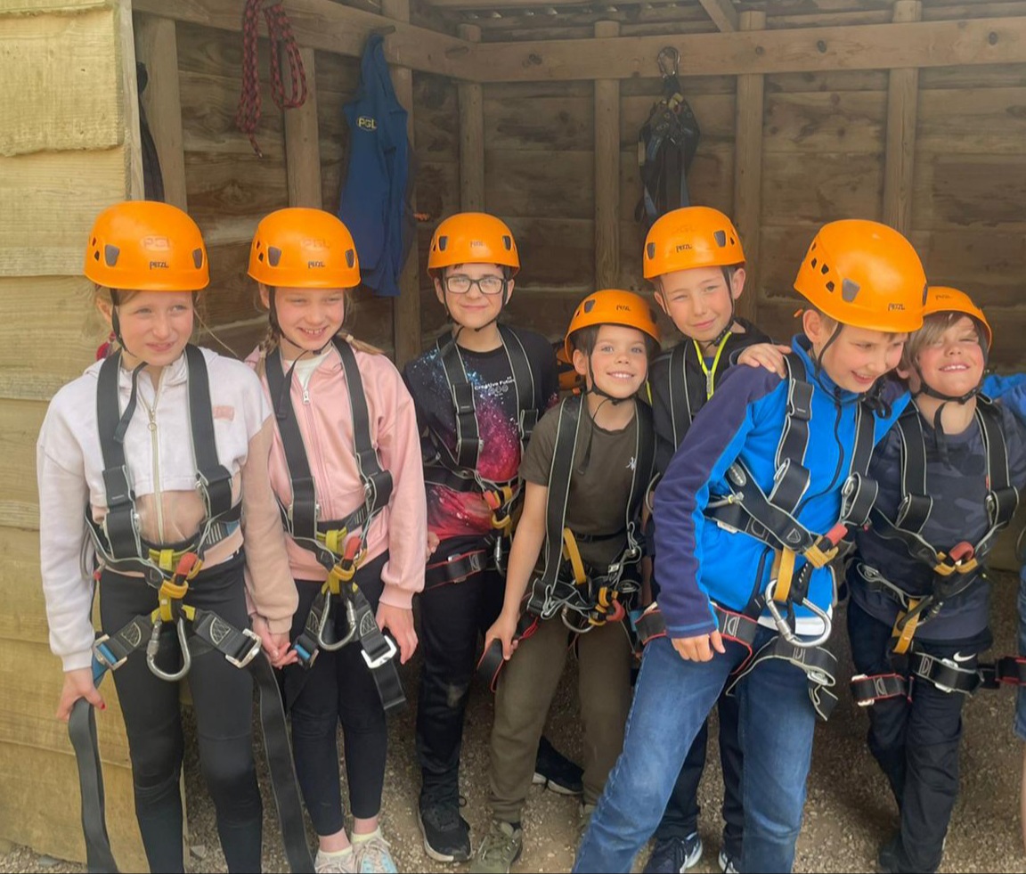 A group of Year 4 children stand ready for a climbing activity, they are wearing safety harnesses and helmets.