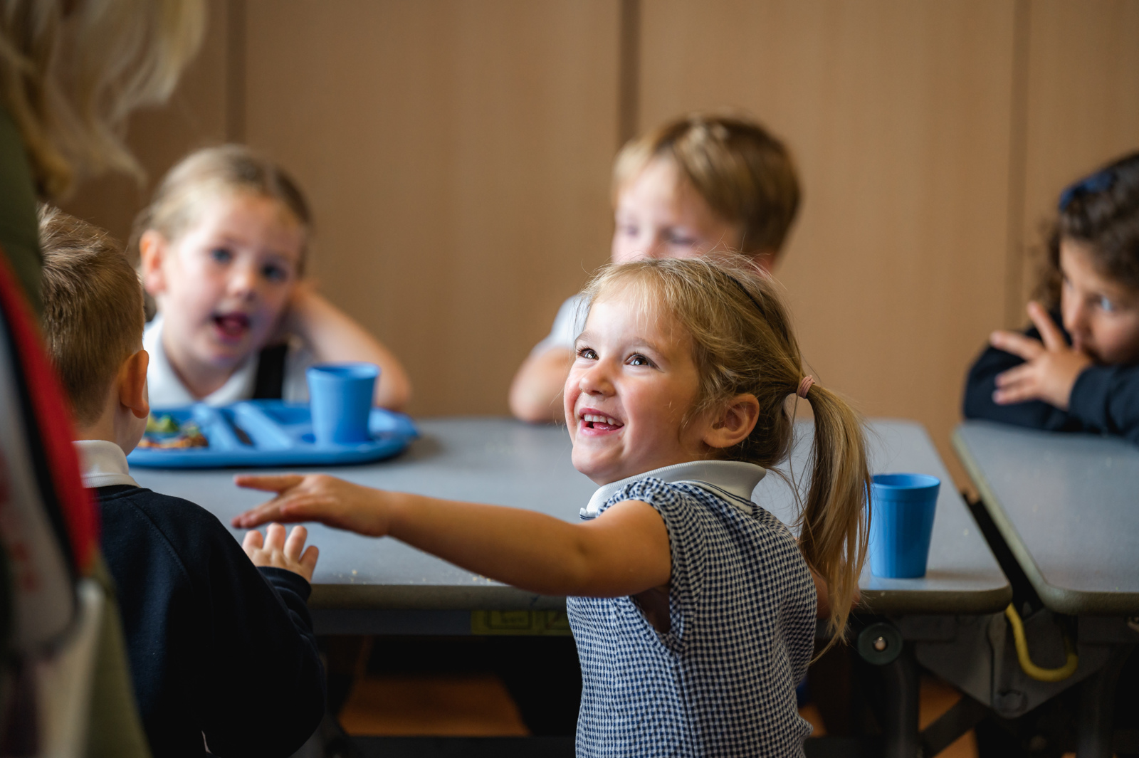 A young girl smiles as she talks to an adult whilst sitting in the dining hall at lunchtime