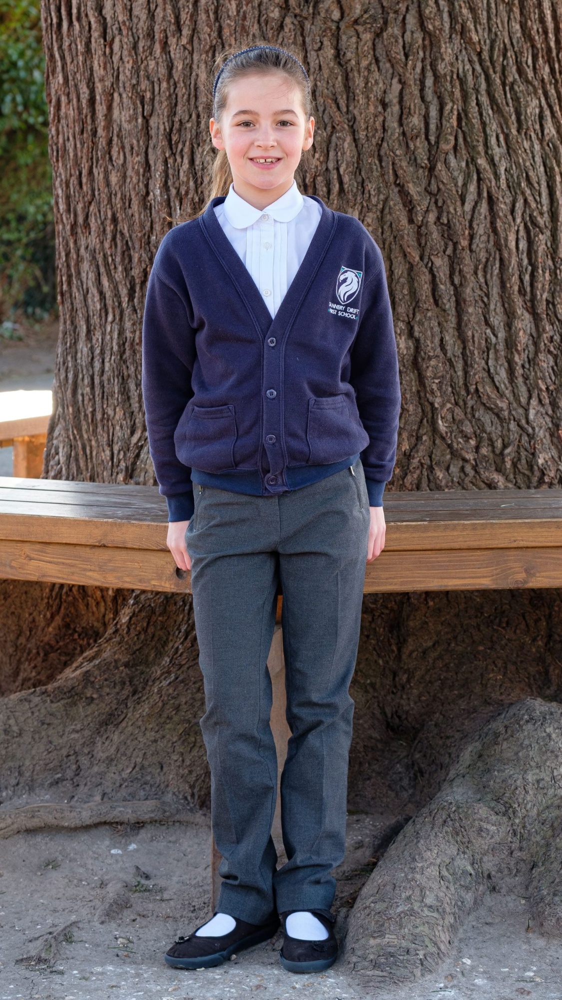 A female pupil stands outside wearing her school uniform, consisting of trousers, a white blouse and a school cardigan