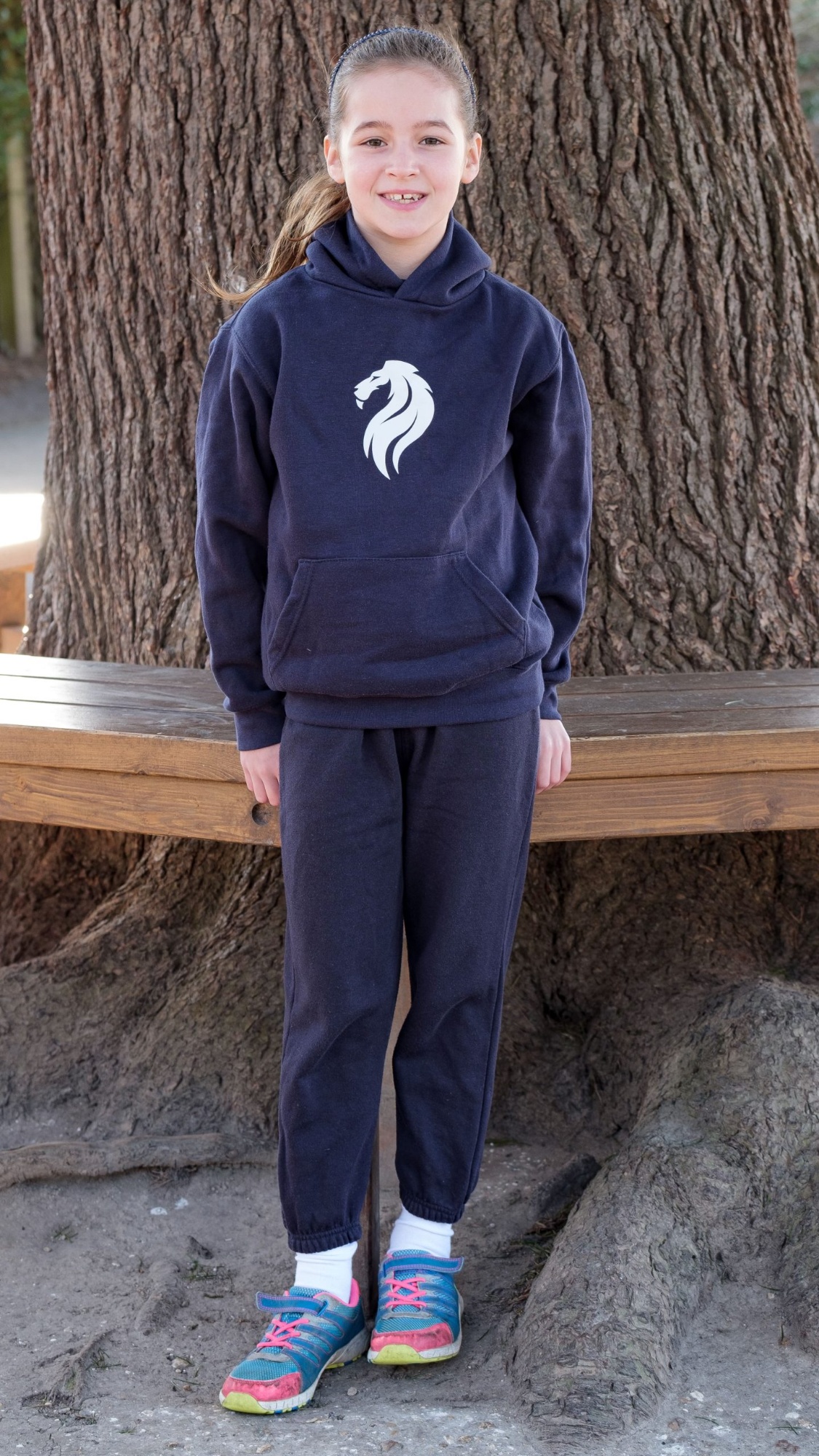 A female pupil stands outside wearing her outdoor PE kit, consisting of a school sports hooded top and jogging bottoms