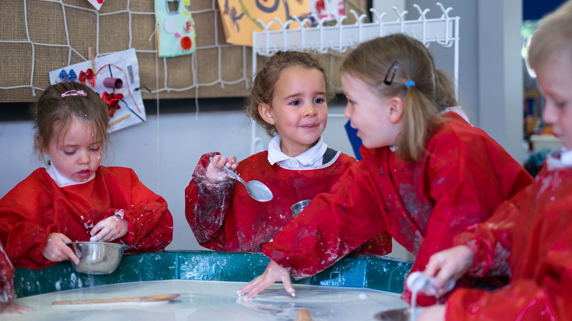 Three girls in the Reception class are wearing aprons while they engage in messy play, using water and kitchen utensils