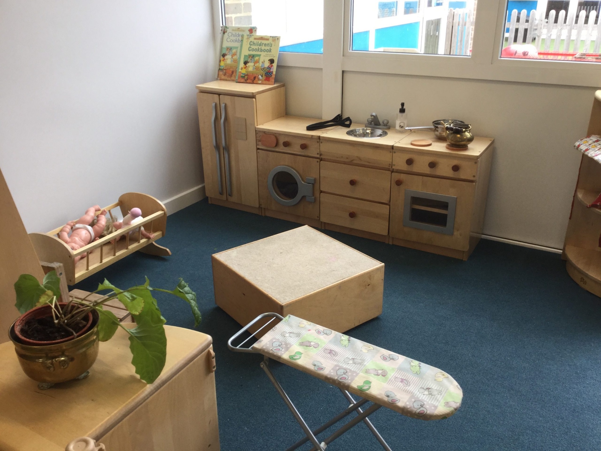 Nursery role play area set up for 'home' role play, with wooden kitchen units, and ironing board and rocking crib with dolls