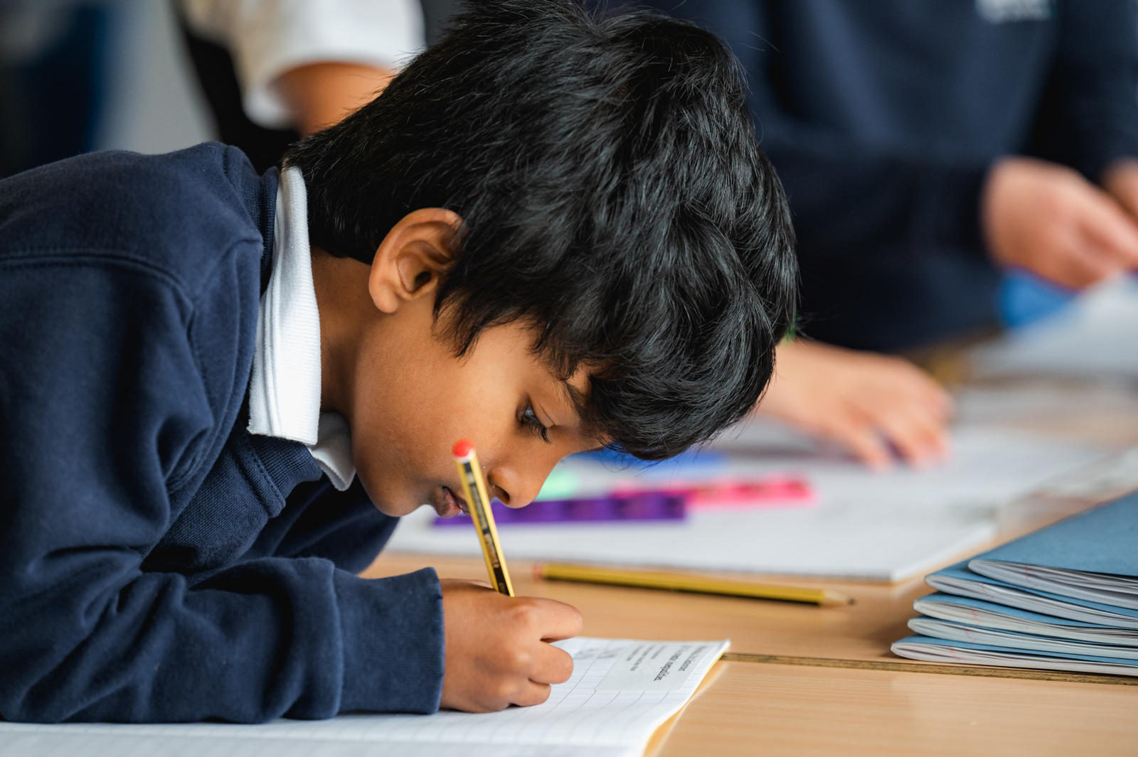 A male pupil writes in his maths book while children in the background use Numicon resources to assist them with their maths task.