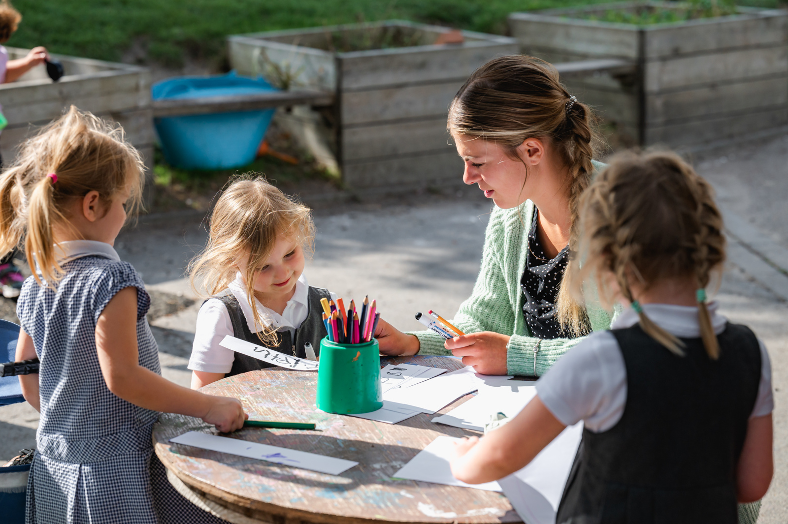 A member of staff sits outside with Reception children teaching them to  write their names