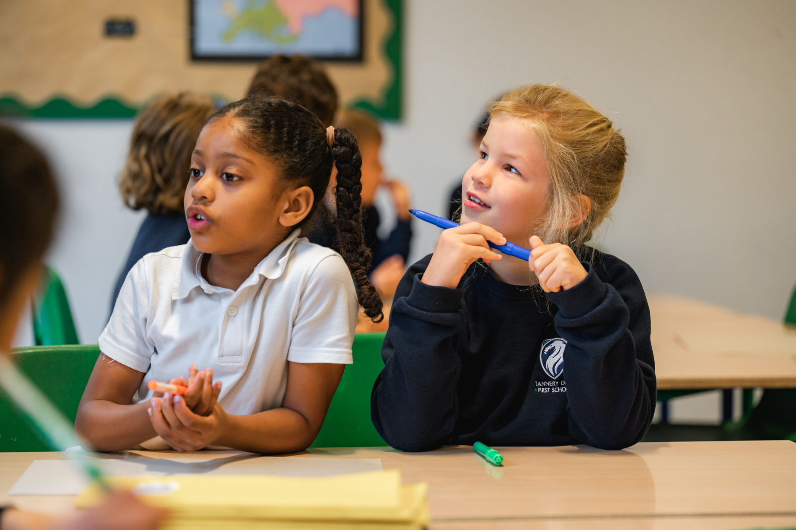 two girls sit next to each other looking at the teacher, one is responding to a question posed by the teacher