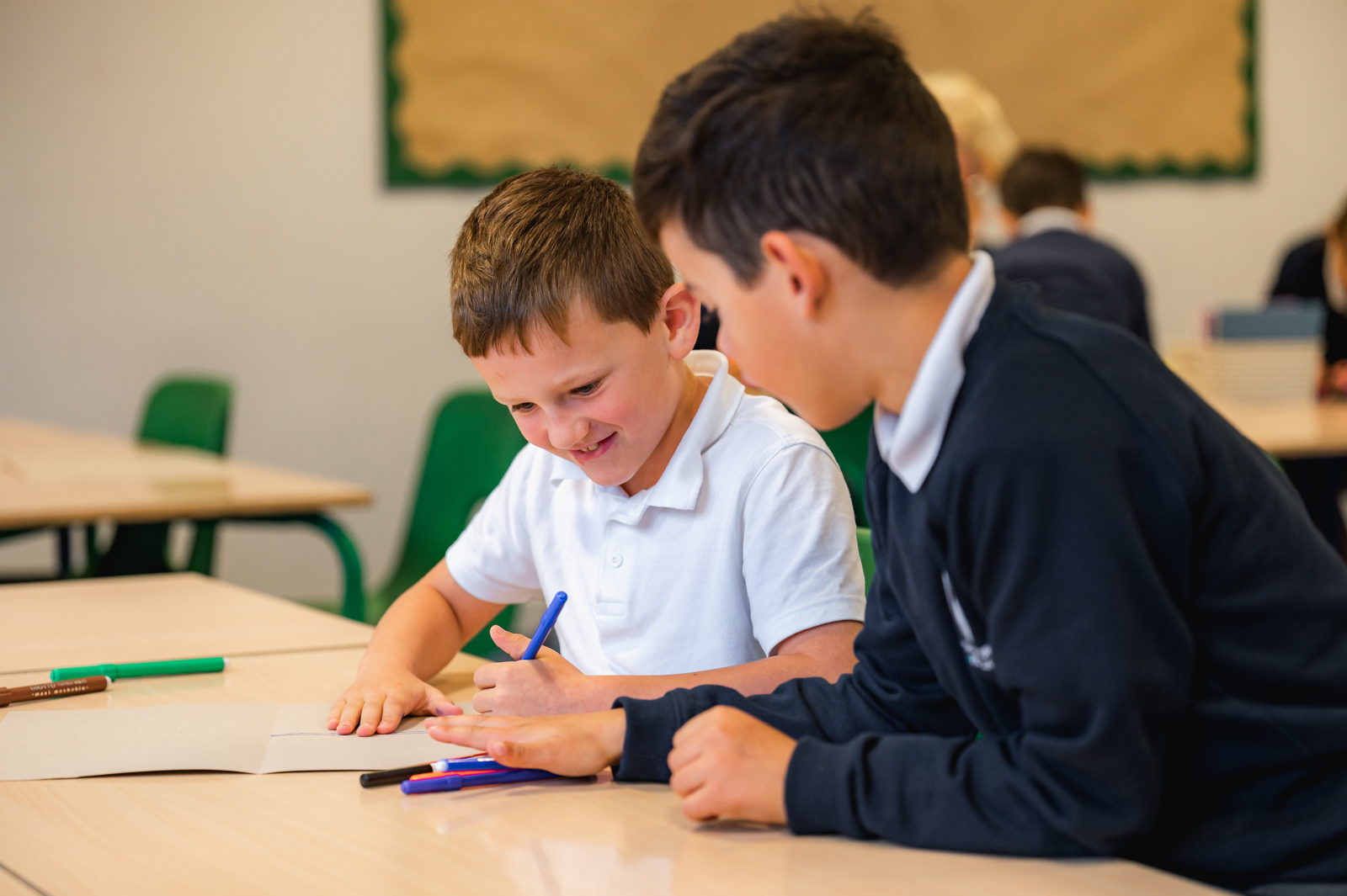 Two boys sit next to each other, working together and using felt tip pens to create a poster 