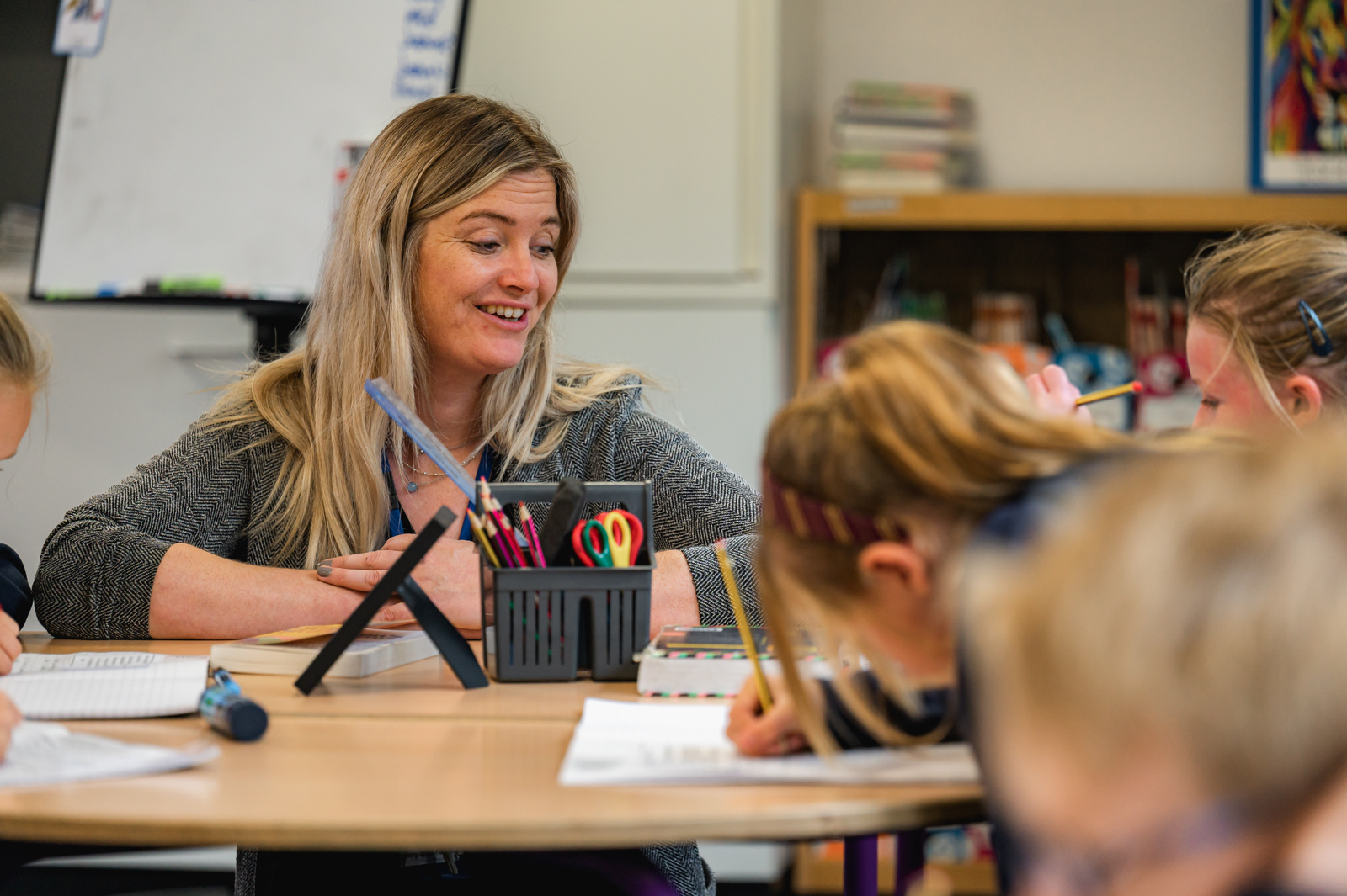 The class teacher sits next to a table talking to the children about the piece of work they are completing