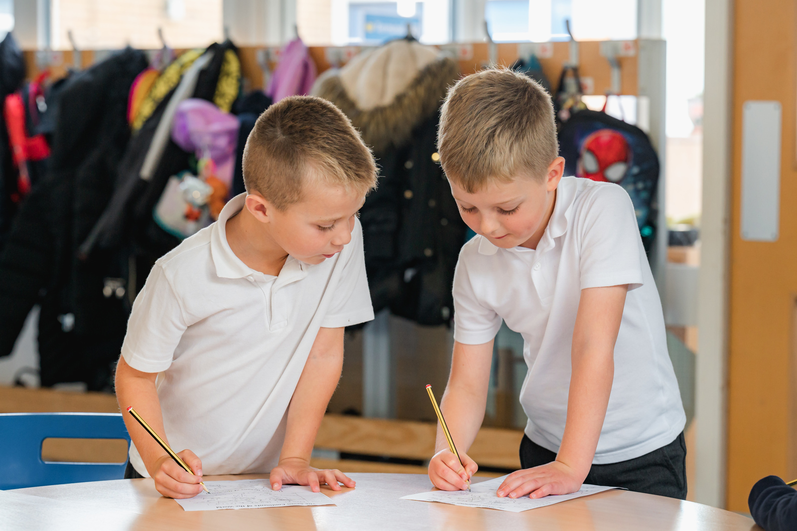 Two boys stand next to each other, each with pencil and paper, working on a learning task together